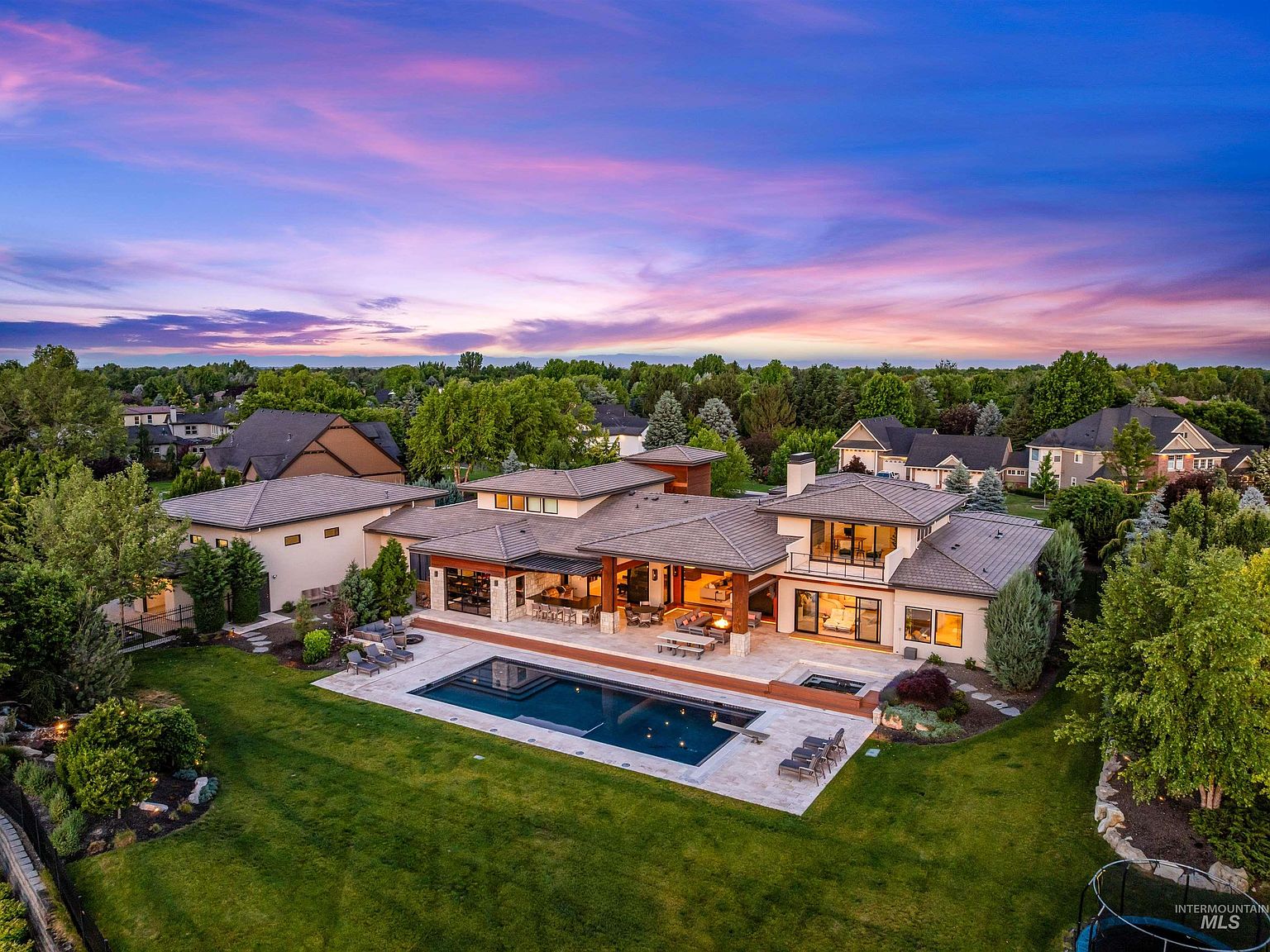 Aerial twilight view of the rear of the estate, the great room glowing and the rectangular pool and spa lit on the travertine terrace below a pink and blue sunset