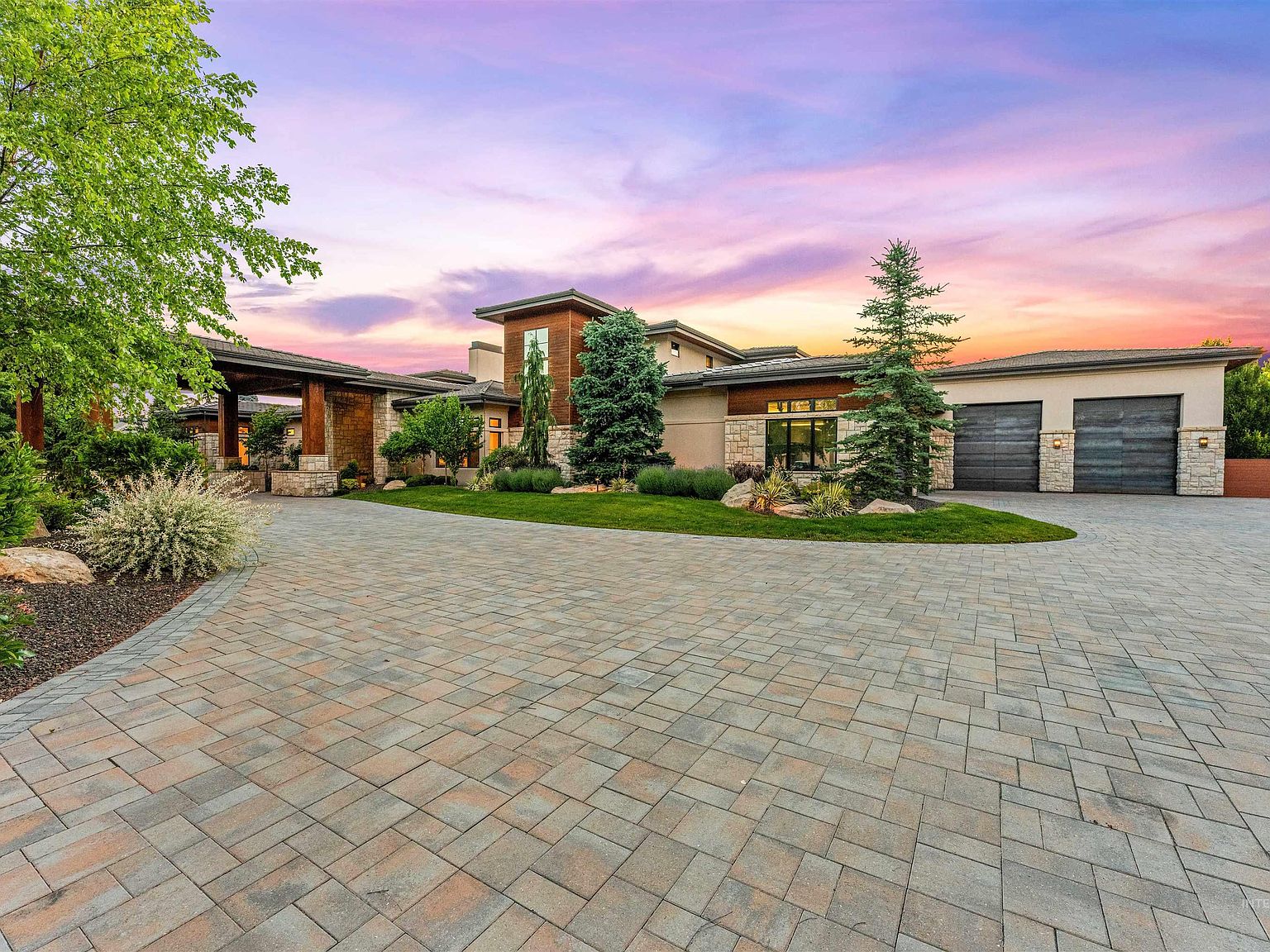Twilight front view of the estate, stone and walnut facade lit warm against a vivid purple and orange sunset sky, with the paver motor court and a two-bay garage