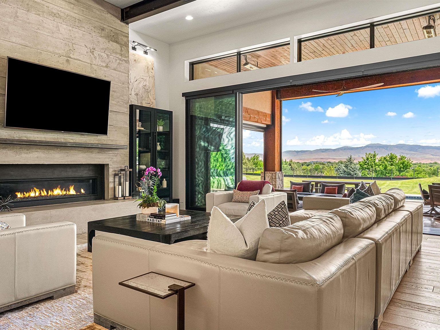 Great room with a concrete-and-walnut linear fireplace and wall-mounted television, the stacking glass wall fully open to a blue-sky mountain view beyond