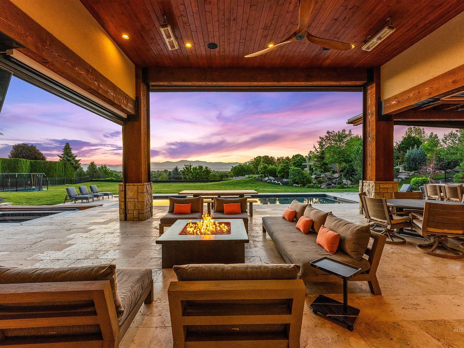 Covered outdoor living room at dusk with a lit fire table, deep sofas, a wood-plank ceiling, and the pool and foothills framed beyond the open beams