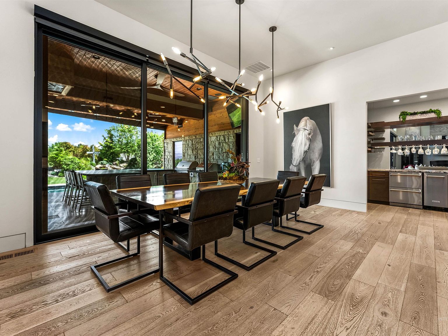 Dining area with a live-edge wood table and black leather chairs under a linear chandelier, the glass wall open to the covered patio and a wet bar to the right