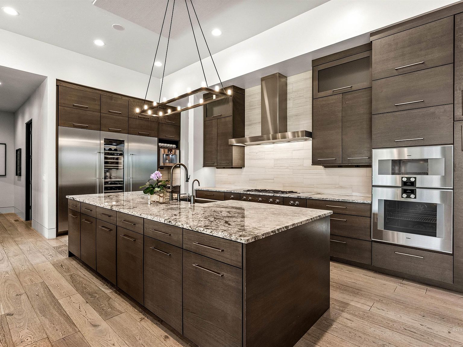 Kitchen with a single quartzite-topped walnut island, a stainless steel range hood over the cooktop, double stainless wall ovens, built-in refrigeration flanked by glass wine columns, and wide-plank hardwood floors