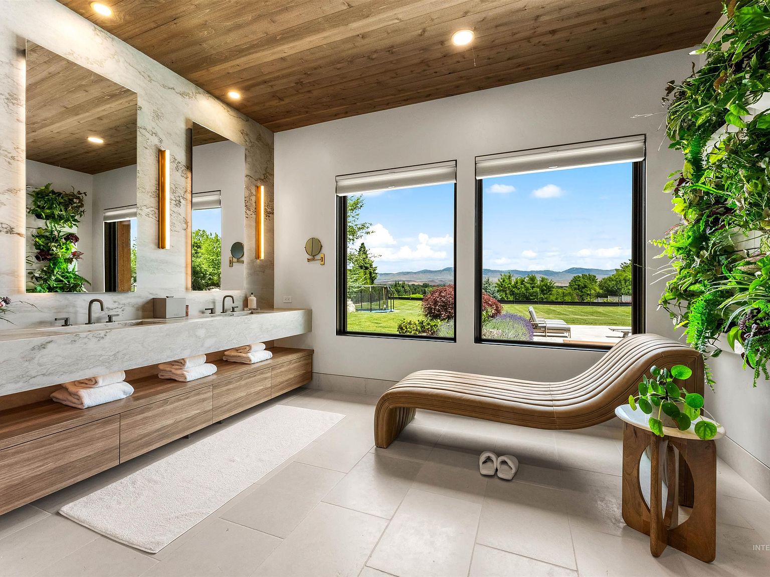 Primary bathroom with a floating quartzite double vanity, full-height stone slab walls, a wood ceiling, a sculptural lounge chair, and windows framing the green lawn and foothills