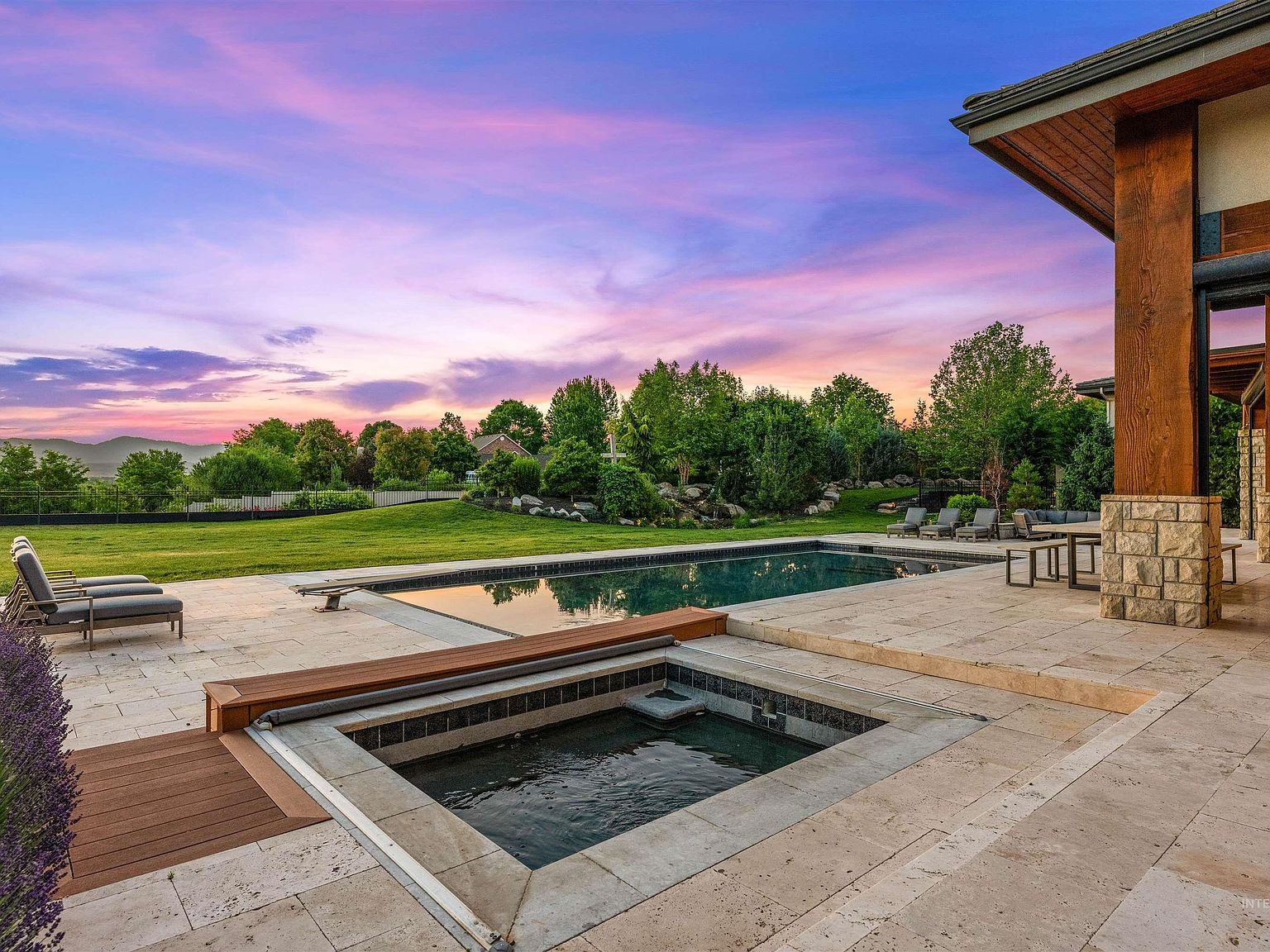 Twilight backyard view of the rectangular pool and the built-in spa with its folding wood cover, set into the travertine deck below a vivid pink and purple sunset over the foothills