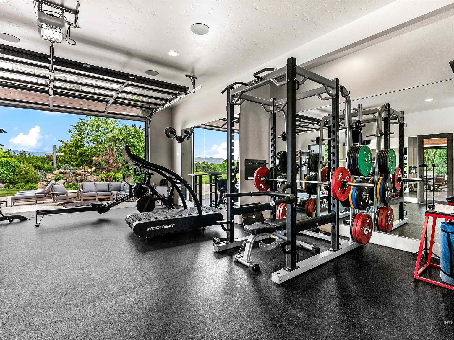 Home gym with a squat rack and color-coded weight plates and a Woodway treadmill, a glass garage door rolled up to open the room onto the patio and lawn