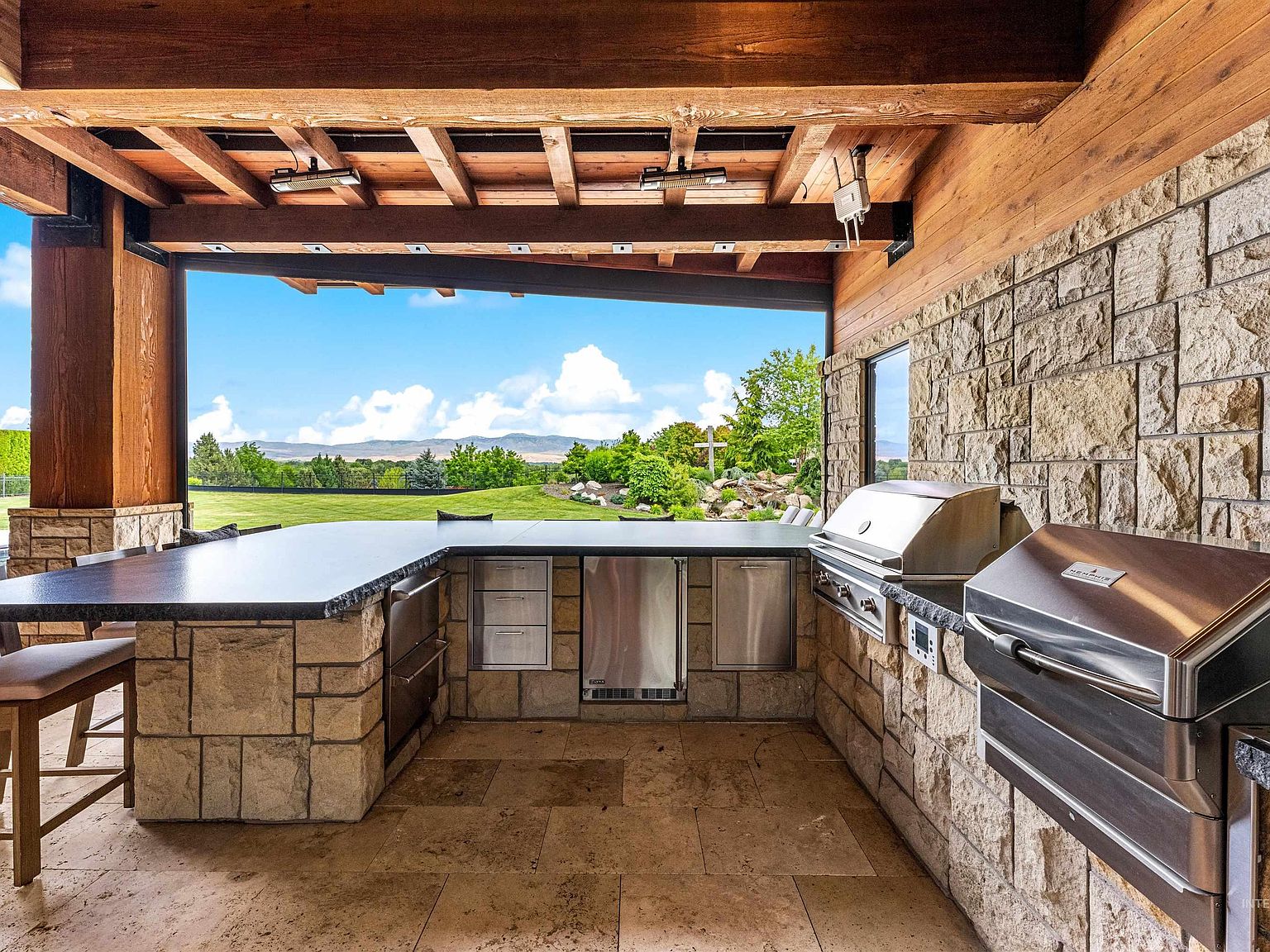 Covered outdoor kitchen in stacked stone with a built-in grill, side burners, and bar seating, open to the lawn and foothills under a beamed wood ceiling