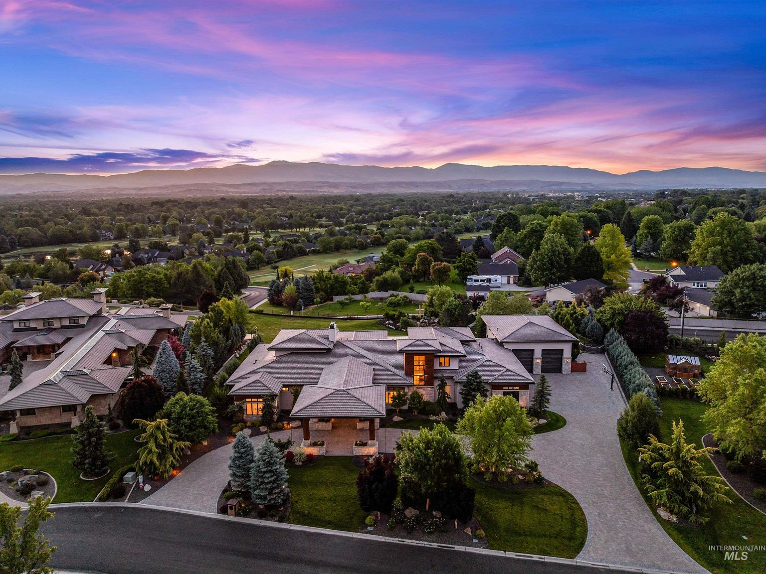 Aerial twilight view of 2748 S Pasa Tiempo Way, a modern tile-roofed estate glowing against the Boise foothills under a pink and gold sunset in Eagle, Idaho